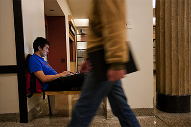 A student sitting in the background in a chair with a laptop. Another students in the foreground walking by with books in his hands.